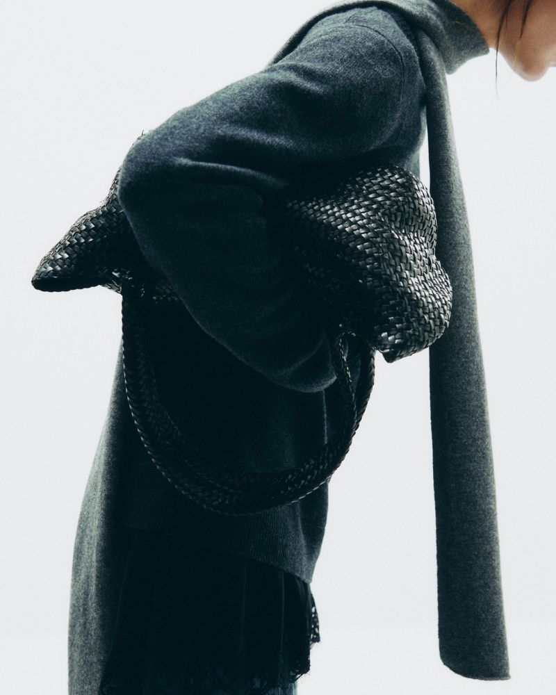 Woman in grey sweater and scarf holds a woven black bag in a studio shot with bright, neutral lighting and minimal background.
