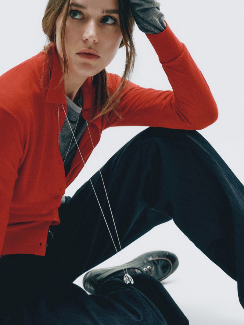 A young adult woman in a red cardigan and dark pants is sitting on the floor with a light background, looking off to the side.