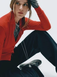 Vivienne Rohner wearing a red button-up knit shirt and wide-leg dark trousers in a seated pose against a white background.