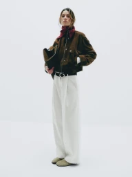 Model wearing a brown suede jacket, burgundy scarf, and wide trousers standing against a seamless white studio backdrop.