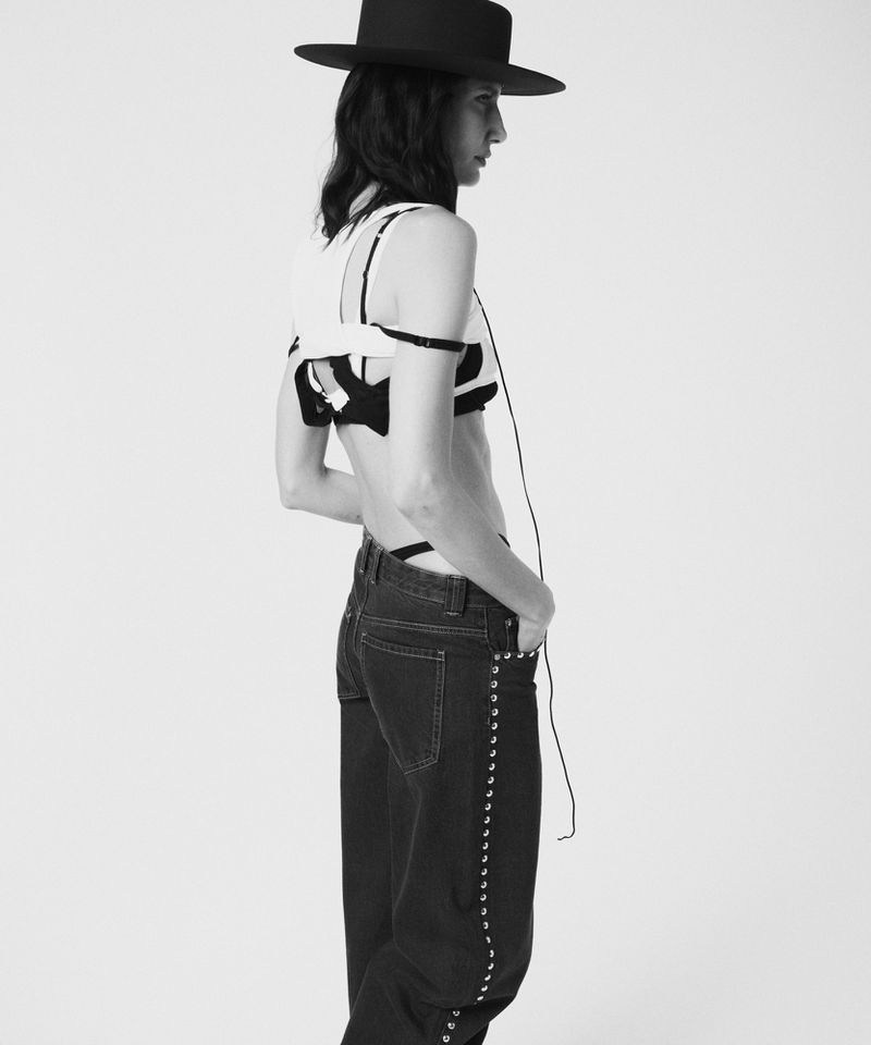 Black and white fashion shot of a slender person with dark hair wearing a hat, modern top, and jeans, posing against a bright, neutral backdrop.