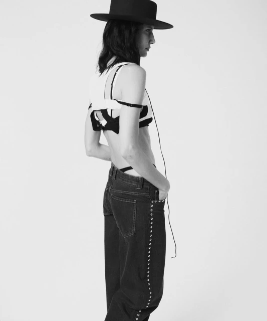 Black and white fashion shot of a slender person with dark hair wearing a hat, modern top, and jeans, posing against a bright, neutral backdrop.