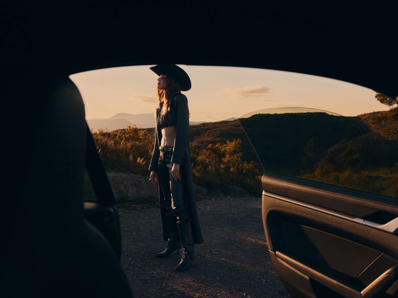 Woman in black bra top, pants, cowboy hat, and coat stands near a car on a rural hillside road during sunset.