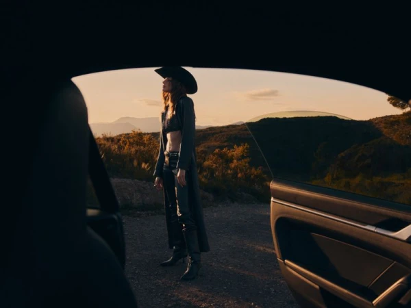 Woman in black bra top, pants, cowboy hat, and coat stands near a car on a rural hillside road during sunset.