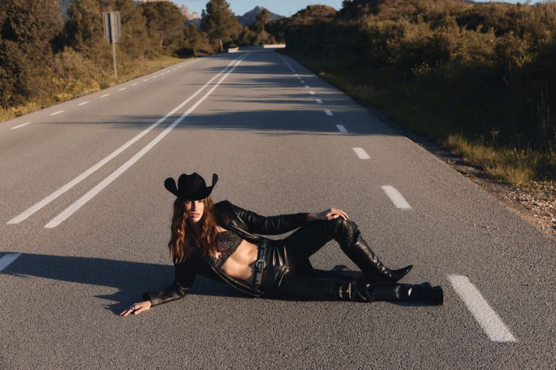 A woman in a cowboy hat and leather outfit poses on a rural road, with trees and hills visible in the distance under a blue sky.