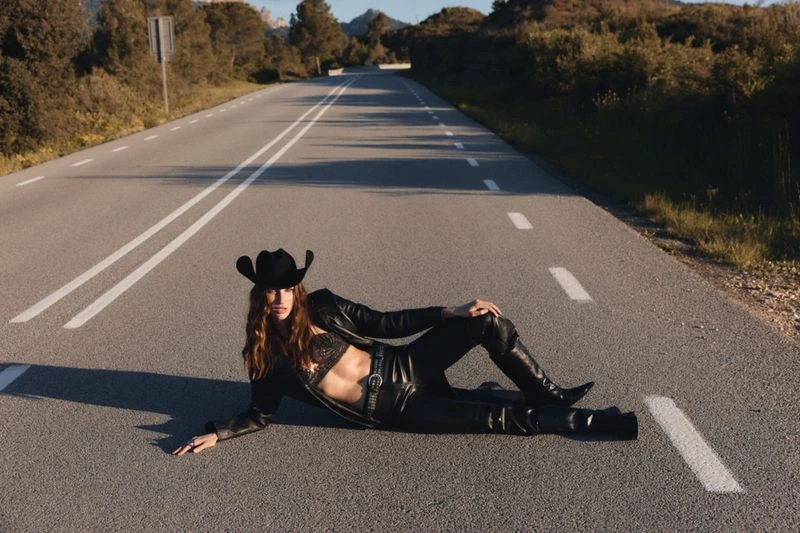 A woman in a cowboy hat and leather outfit poses on a rural road, with trees and hills visible in the distance under a blue sky.
