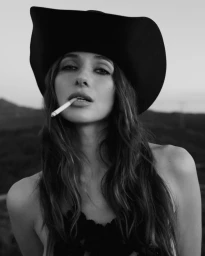 Monochrome portrait of a woman wearing a wide brim hat holding a cigarette in her mouth against an outdoor horizon.