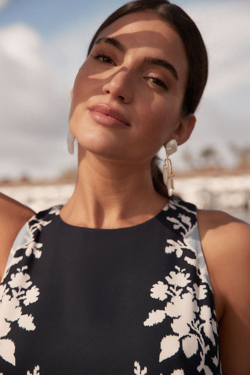 A woman poses in a sleeveless black dress with a floral pattern, complemented by dangling earrings and natural outdoor lighting.