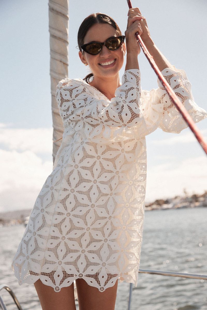 Woman in white lace dress and sunglasses holding onto a boat rigging, smiling on a bright sunny day in a nautical setting.