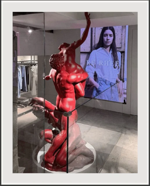 Sculpture of two figures in red inside retail store behind glass with an advertisement in background featuring a woman in casual wear.
