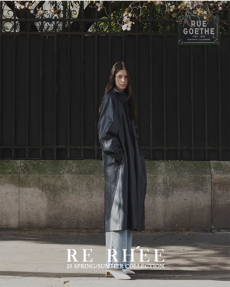 A slender woman in a dark coat and light denim stands in an urban outdoor setting, framed by an iron fence and a Parisian street sign.