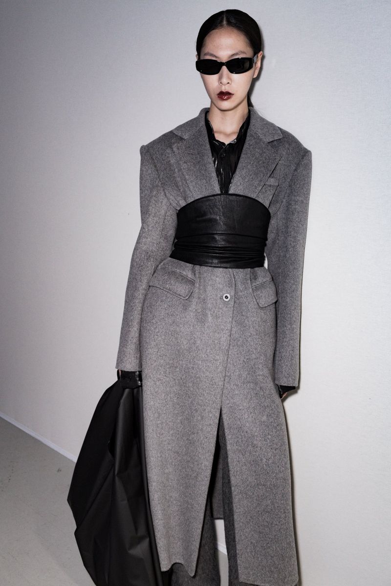 A stylish Asian woman in a grey wool coat and black leather corset poses against a light wall, wearing sunglasses and black gloves.