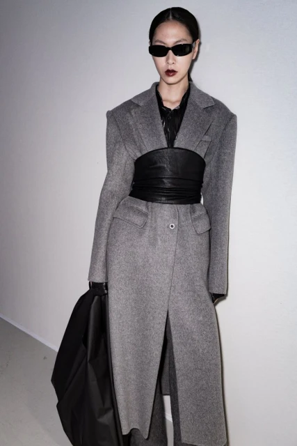 A stylish Asian woman in a grey wool coat and black leather corset poses against a light wall, wearing sunglasses and black gloves.
