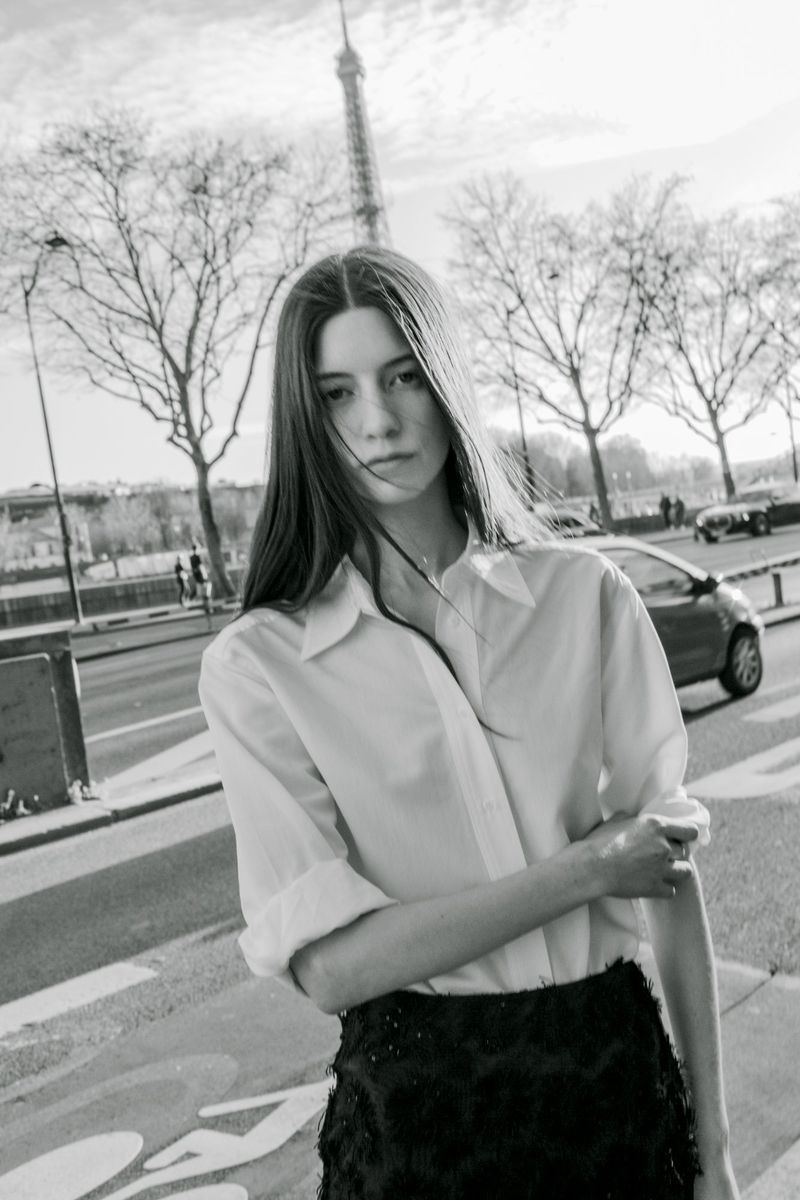 A woman in a white shirt poses in Paris, France, with the Eiffel Tower visible in the background, captured in black and white.