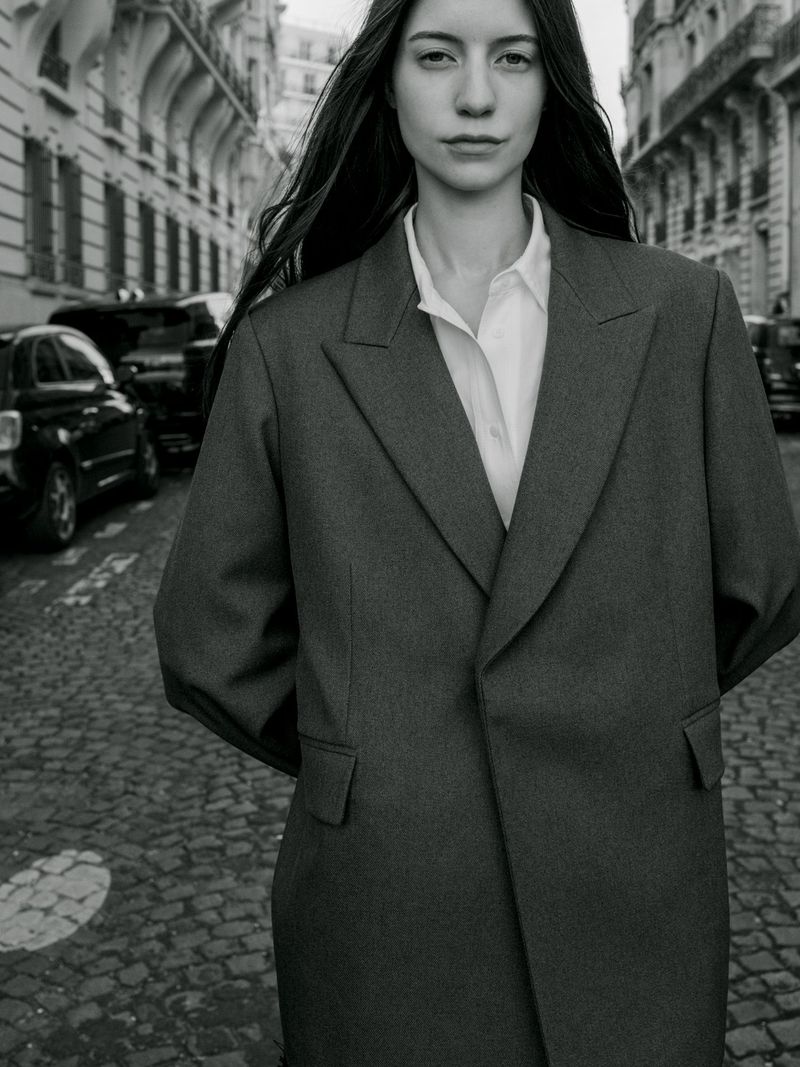 Monochrome portrait of a woman in an oversized tailored coat and white collared shirt standing on a cobblestone street.