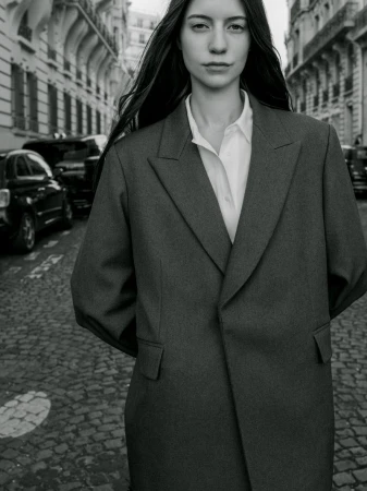 Monochrome portrait of a woman in an oversized tailored coat and white collared shirt standing on a cobblestone street.
