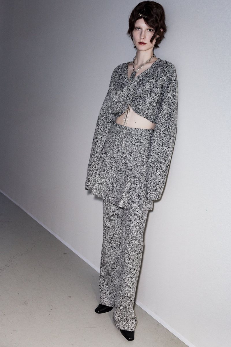 Fashion model wearing a grey speckled knit set posing in a studio against a white wall, showcasing contemporary apparel design.