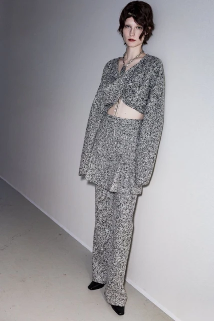 Fashion model wearing a grey speckled knit set posing in a studio against a white wall, showcasing contemporary apparel design.