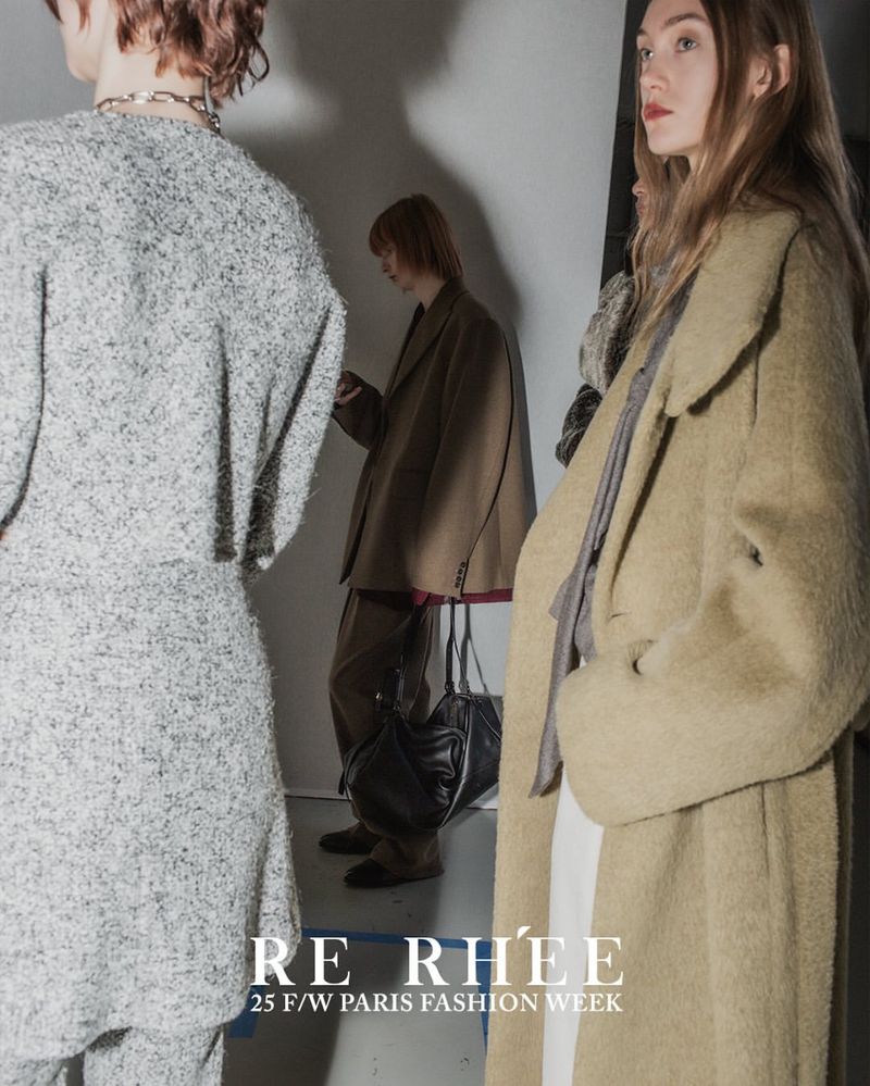 Three fashion models in earth-tone outfits backstage at a show; one in grey textured suit, the other in brown and beige oversized coats.