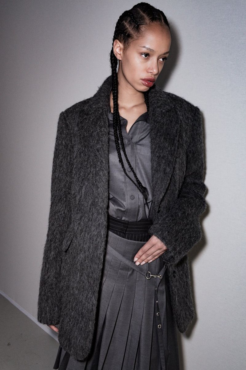 Woman with braided hair in grey mohair blazer, button-down shirt, and pleated mini skirt, posing in an indoor setting with neutral background.
