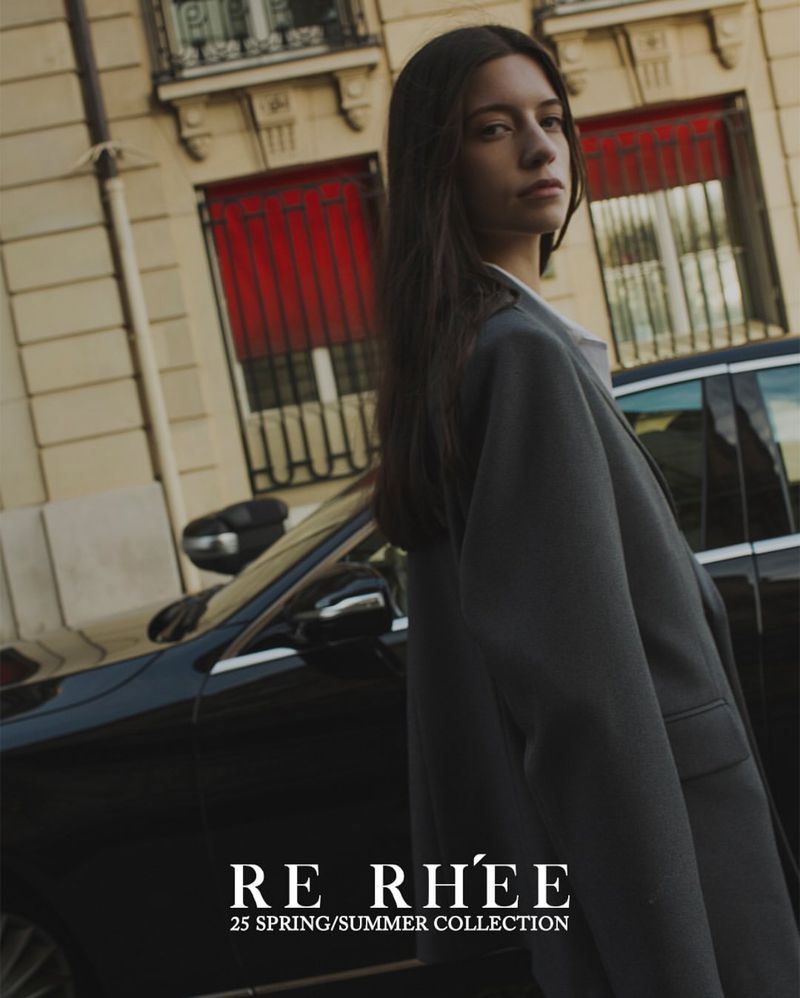 Brunette woman in an oversized grey blazer poses next to a black car on a city street in front of a light-colored building.