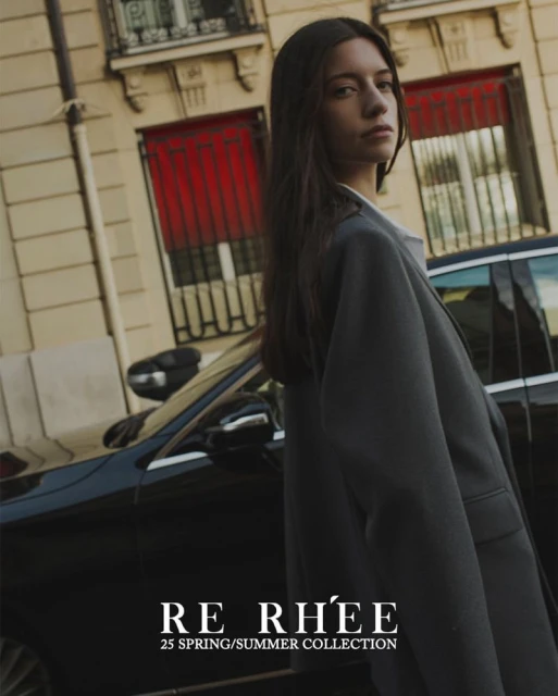 Brunette woman in an oversized grey blazer poses next to a black car on a city street in front of a light-colored building.