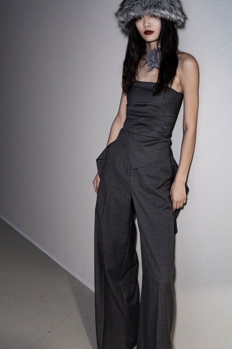 Woman in grey strapless jumpsuit with wide legs and fluffy hat poses against a white wall in a fashion editorial, shot at eye-level.