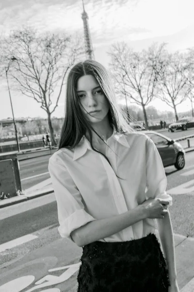 Female model in an open-collar white shirt and dark textured skirt posing on a city street with the Eiffel Tower in the background.