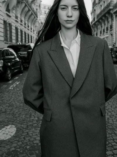 Monochrome portrait of a woman in an oversized tailored coat and white collared shirt standing on a cobblestone street.