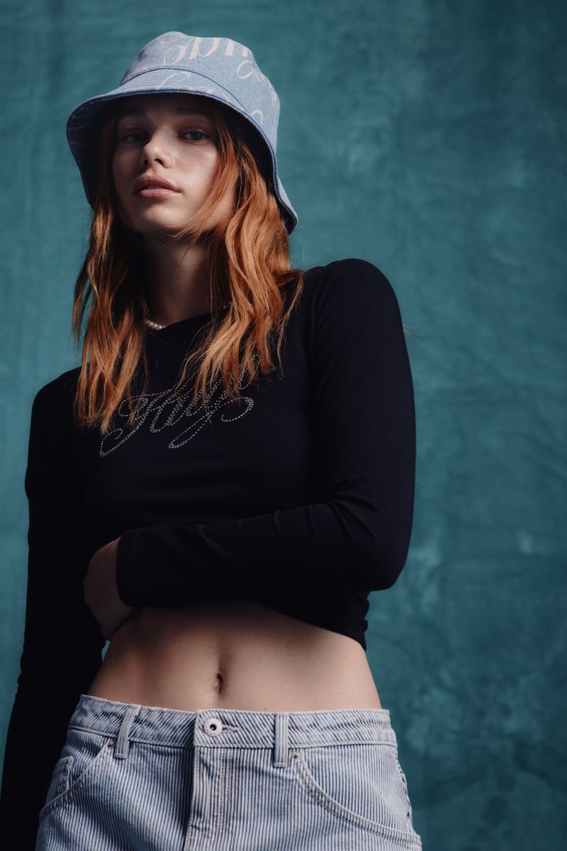 Young woman in a black long-sleeve crop top, striped jeans, and matching bucket hat poses in front of a textured teal backdrop.