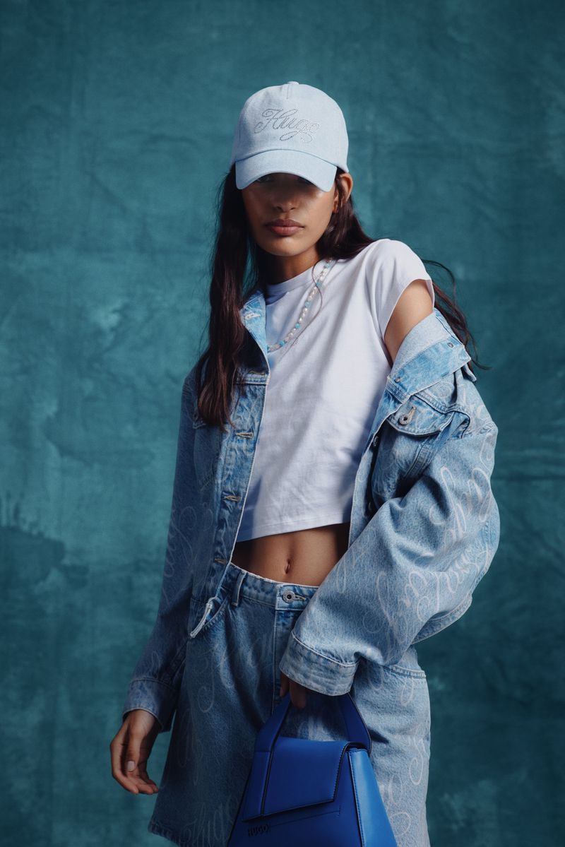 Woman in denim jacket and skirt with white top and blue purse against blue backdrop, posing in a studio shot. Fashion photography.