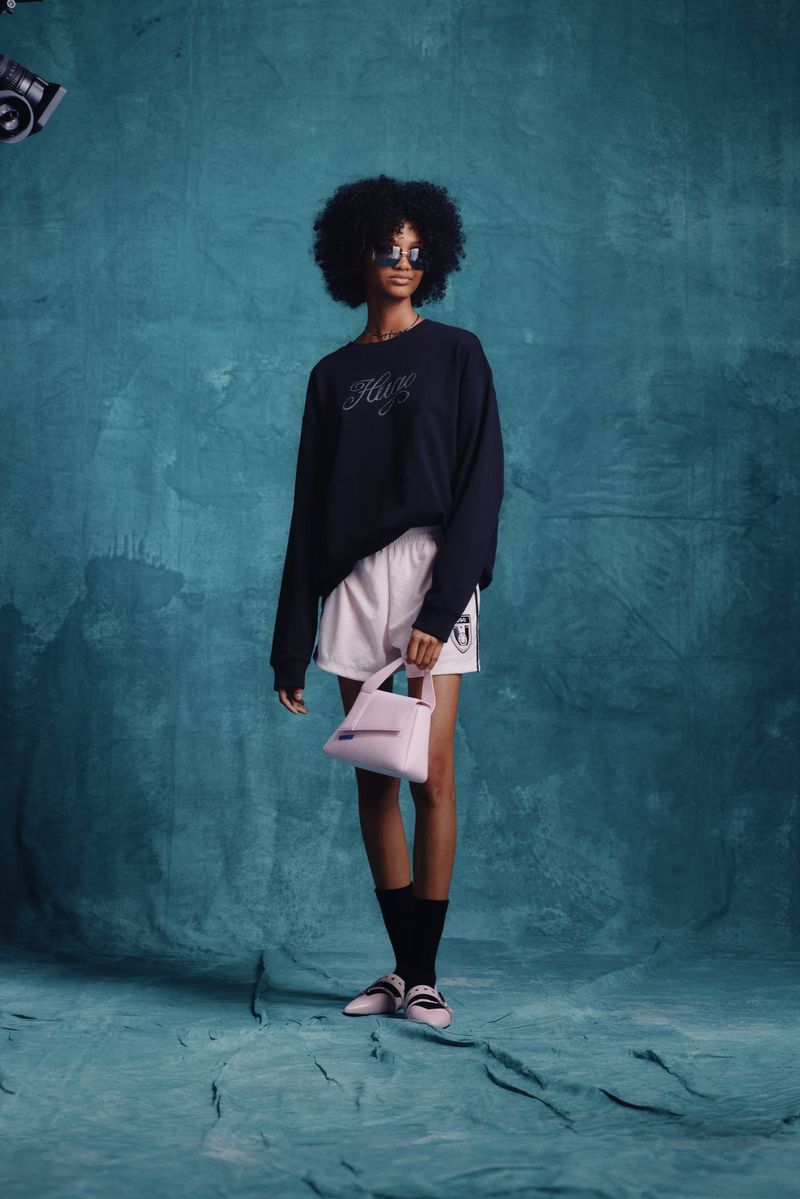 Full-length studio shot of a Black woman wearing a sweatshirt, shorts, socks, and flats, and holding a purse against a teal background.