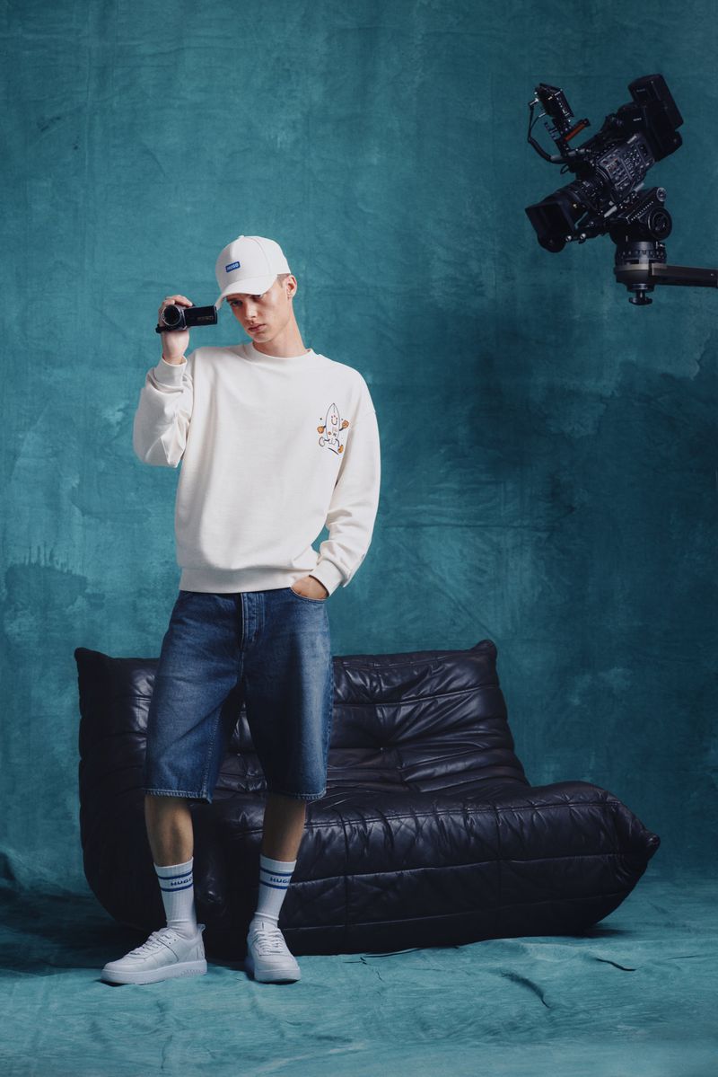 A young adult man in a white sweatshirt and denim shorts holds a camcorder in a photo studio with professional video camera equipment.