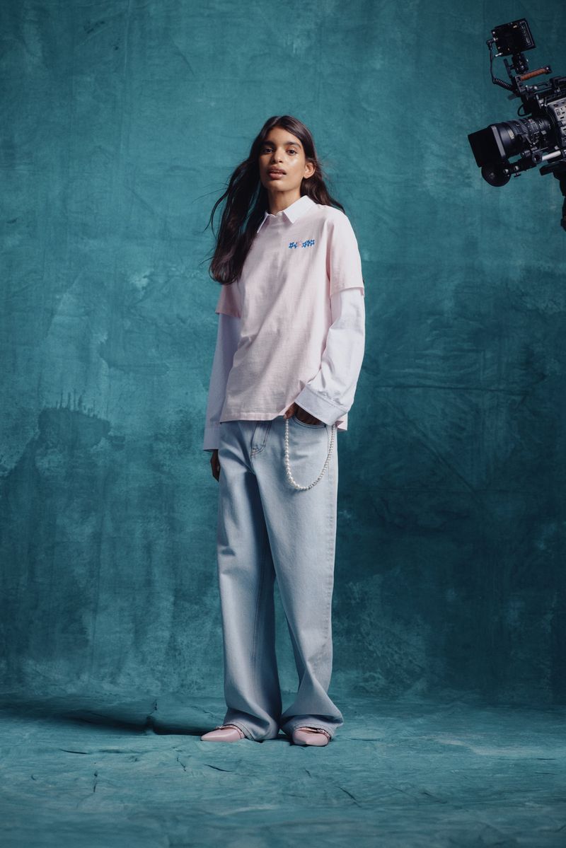 Studio shot of a young adult female model in a pink and white top with wide-leg jeans, posing in front of a textured teal backdrop.
