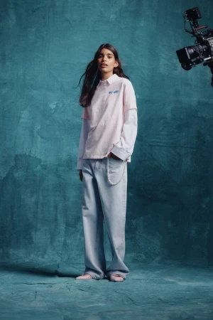 Studio shot of a young adult female model in a pink and white top with wide-leg jeans, posing in front of a textured teal backdrop.