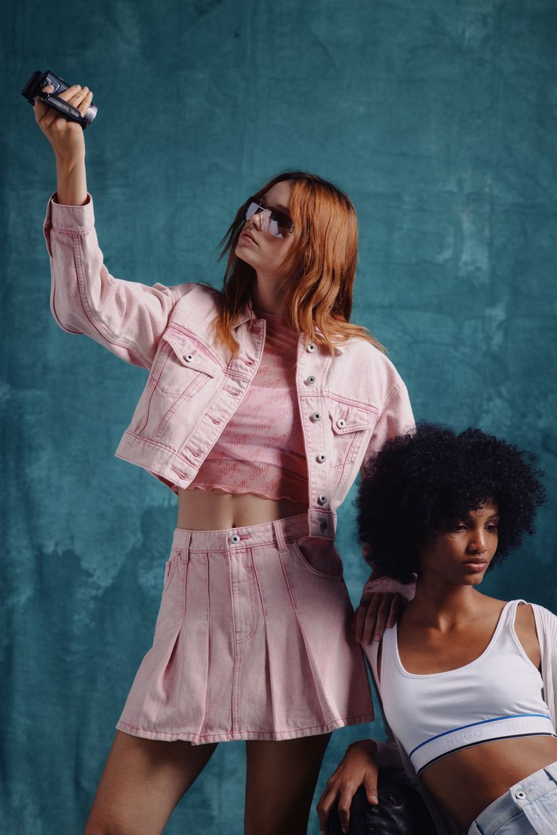 Two women model pink denim jacket and skirt, and a white crop top against a blue textured backdrop in a fashion editorial shot.