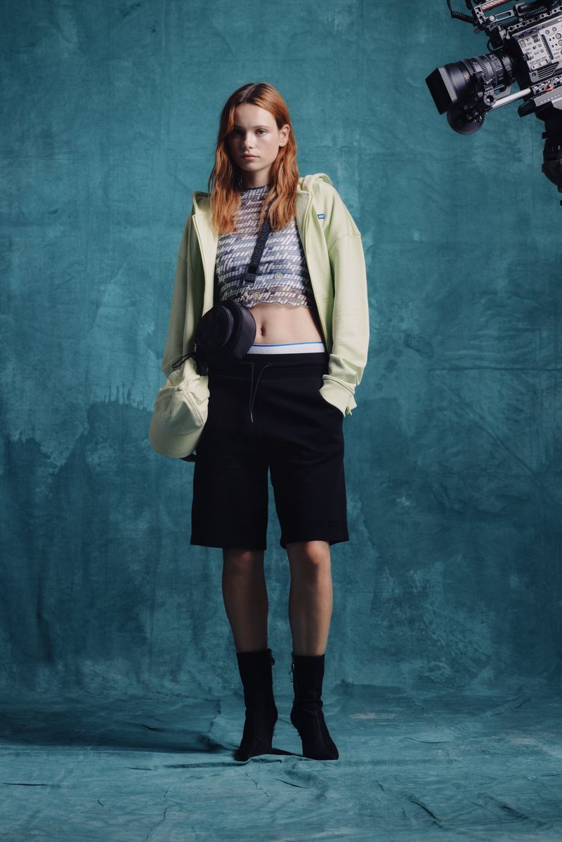 Full-length shot of a red-haired fashion model posing in black shorts, light-green hoodie, and black booties against a teal studio backdrop.