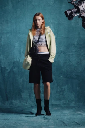 Full-length shot of a red-haired fashion model posing in black shorts, light-green hoodie, and black booties against a teal studio backdrop.