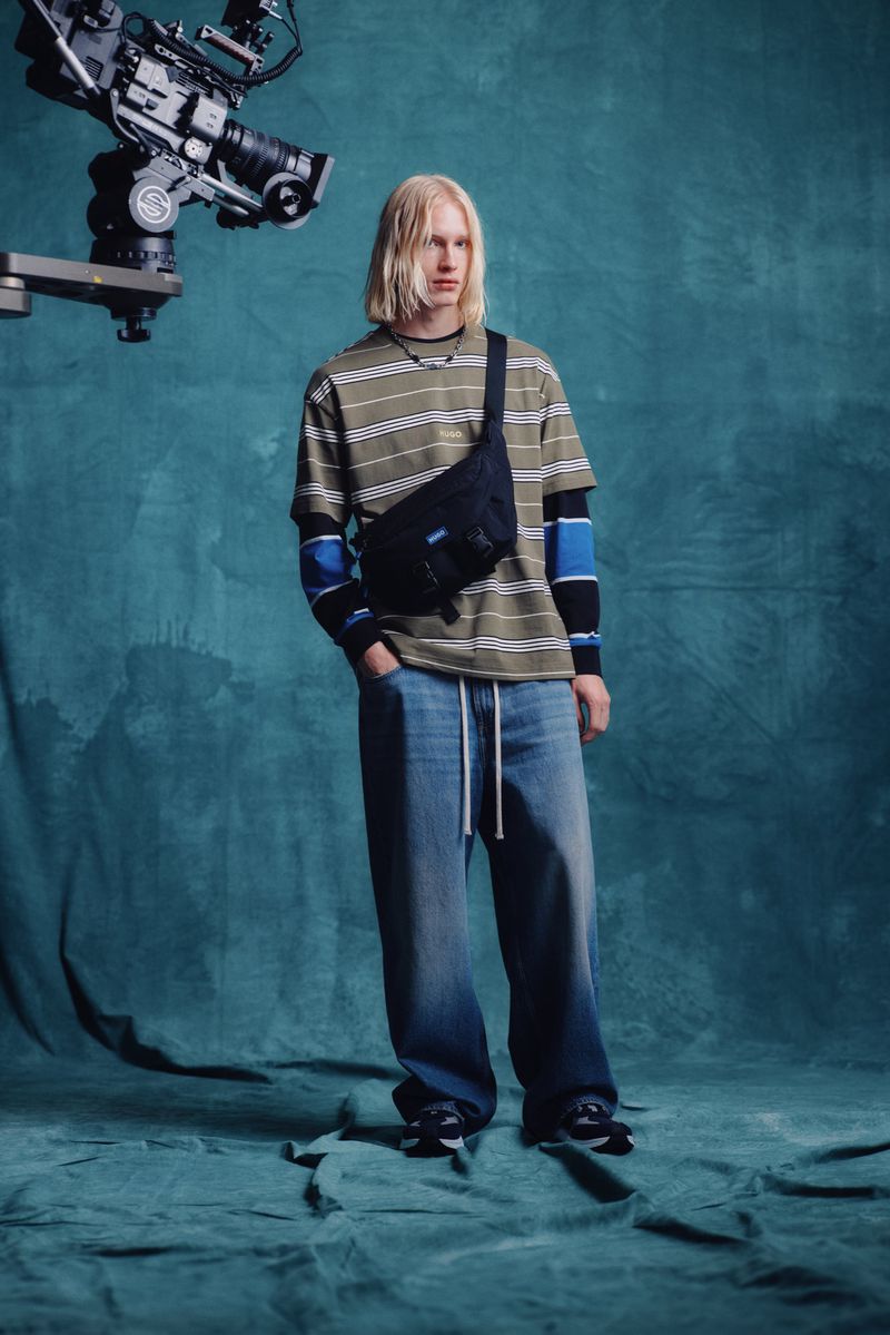 Male fashion model poses in studio with striped shirt, jeans, and fanny pack, alongside a professional film camera on a boom.