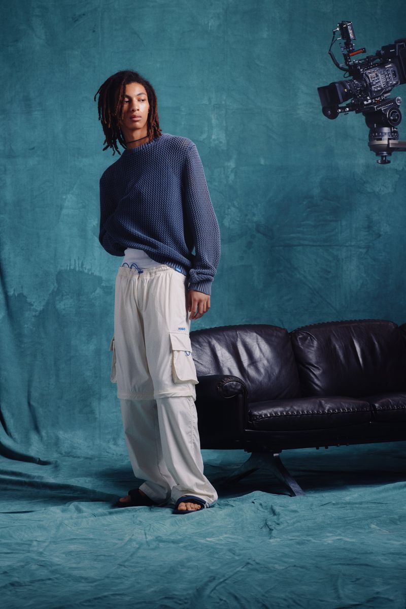 A model wearing a blue sweater and loose white pants poses in a photo studio with a camera, black leather couch, and blue backdrop.