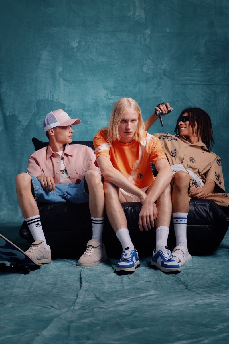 Three young adult male fashion models in colorful streetwear outfits sit on a black leather couch in a photo studio setting.