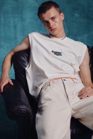 A young man seated in a dark leather armchair wearing an off-white sleeveless graphic top and matching trousers against a teal backdrop.
