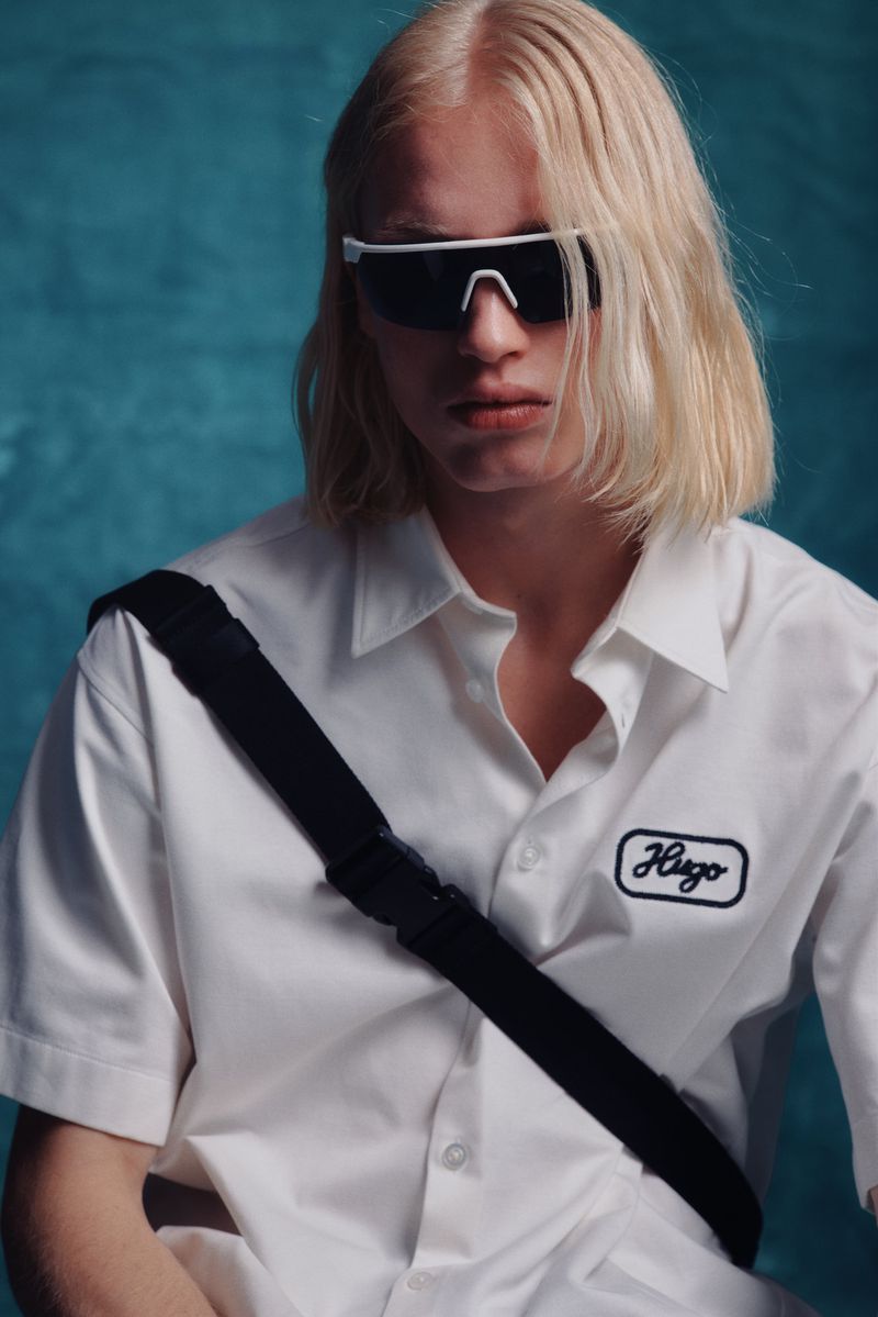 Stylish young adult with blond hair poses in a white Hugo shirt with a black strap and white sunglasses against a blue backdrop.