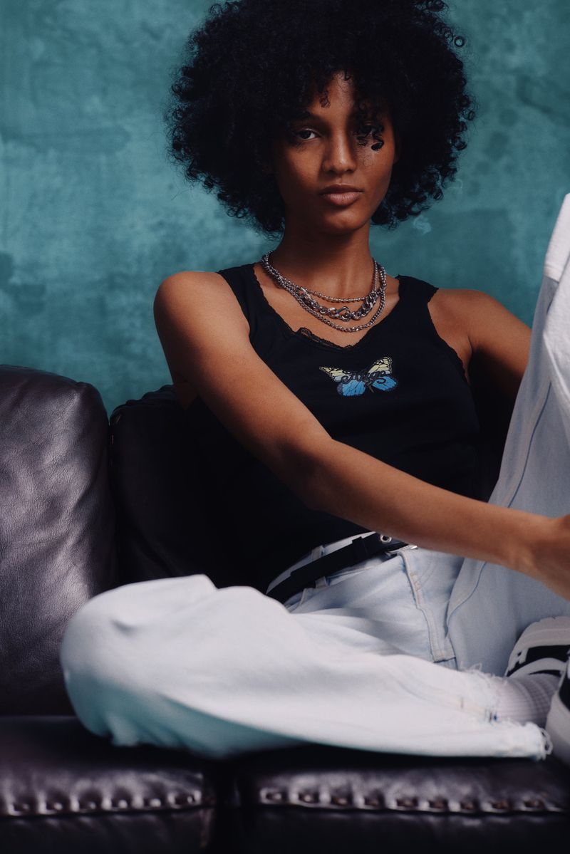 A fashionable young woman with an afro poses on a couch wearing a black top, white pants, and layered silver necklaces.