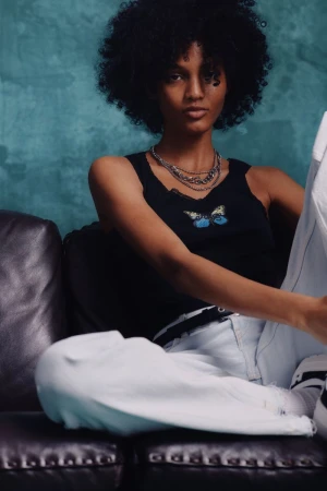 A fashionable young woman with an afro poses on a couch wearing a black top, white pants, and layered silver necklaces.