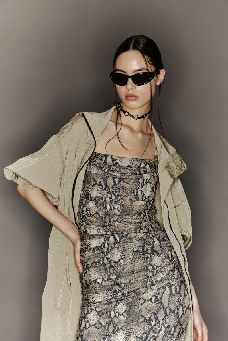 A woman poses in a snake-print dress and sunglasses, wearing a light trench coat against a grey backdrop in a fashion shot.