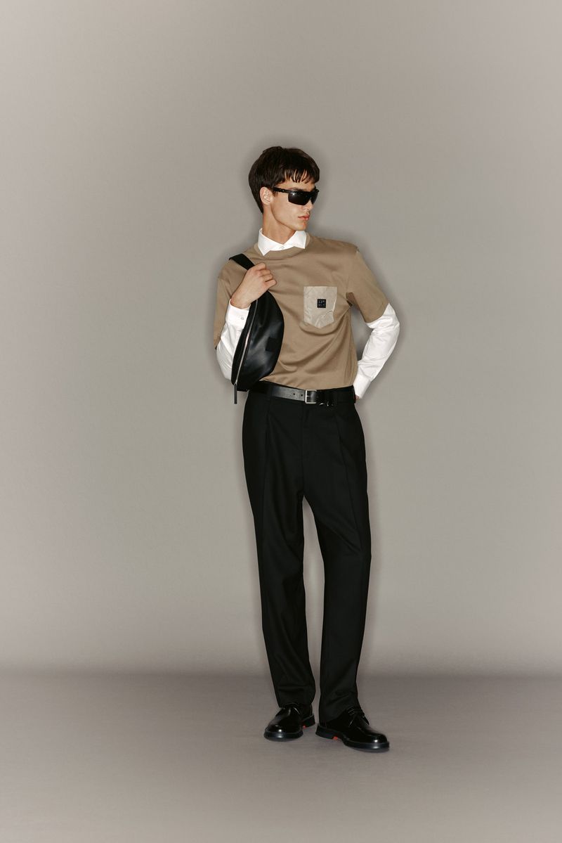 Full-length shot of an adult male model wearing a brown t-shirt with black dress pants, sunglasses, and dress shoes in a studio.