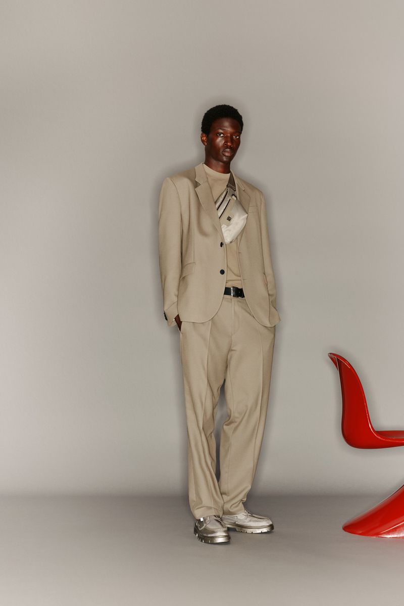 A slender Black man in a beige suit, t-shirt, and silver sneakers poses in a studio with a red plastic chair visible on the right.