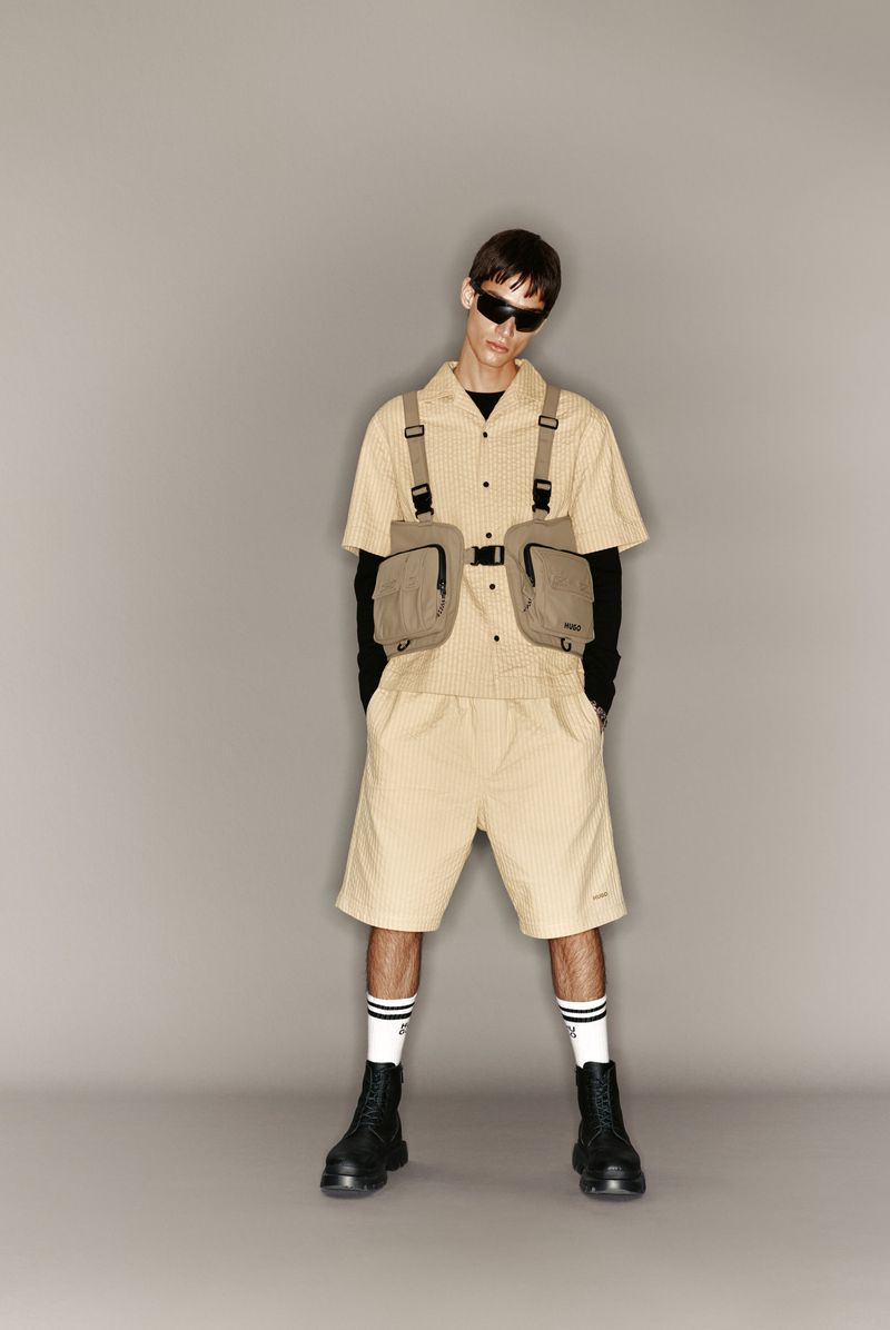 Man in khaki button-down shirt and shorts with vest poses in front of grey background in full shot, accessorized with sunglasses and boots.