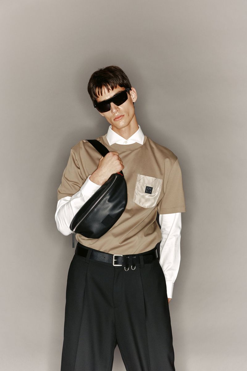 Adult male wearing a beige t-shirt with a light-colored pocket, black pants, and black sunglasses, and a black bag across his chest.